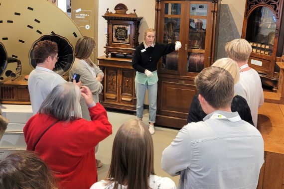 Eine Gruppe von Menschen schaut auf mechanischen Musikautomaten, welche von einer Führungsperson erklärt werden.